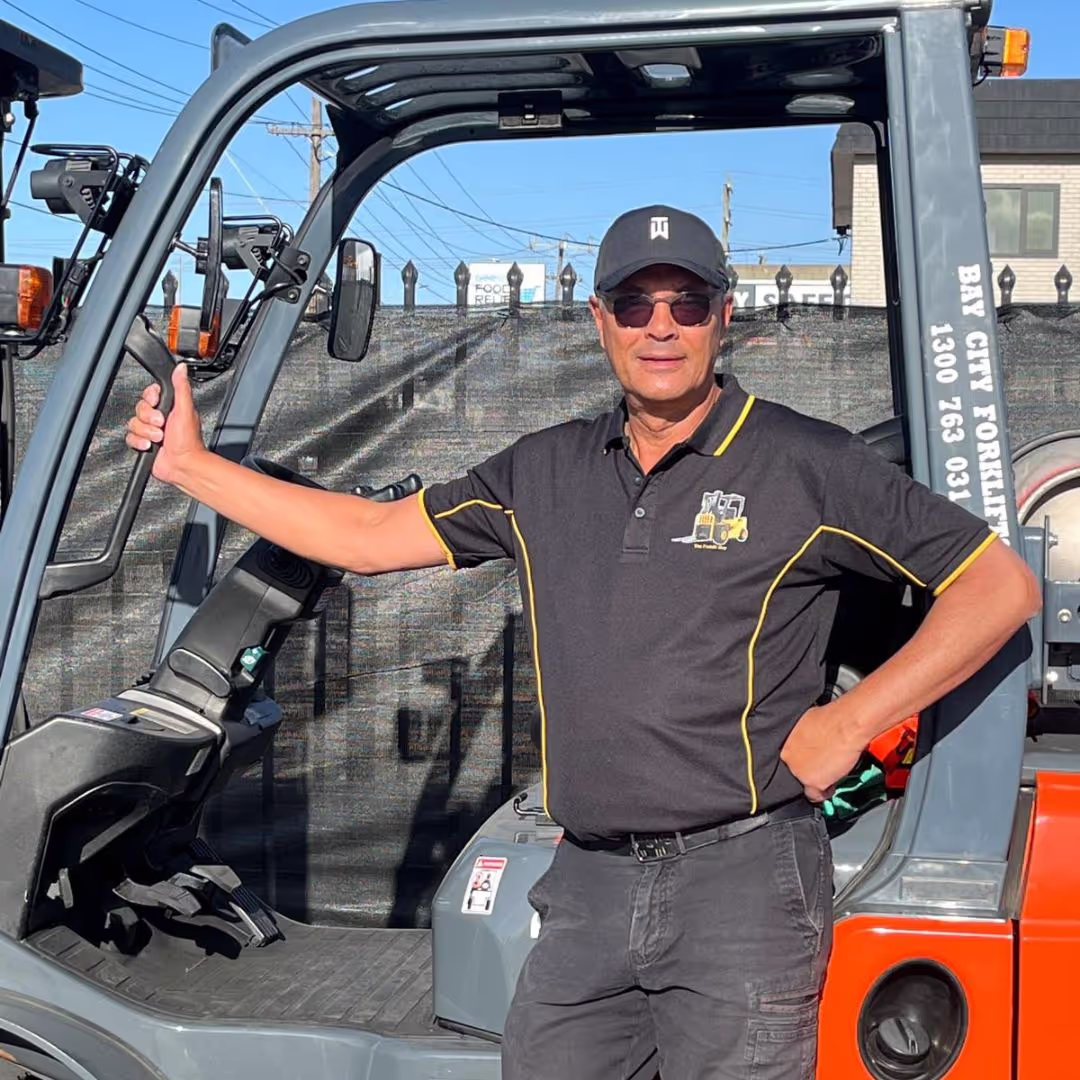 Man wearing sunglasses and a cap stands with one hand on the frame of a forklift and the other on his hip.